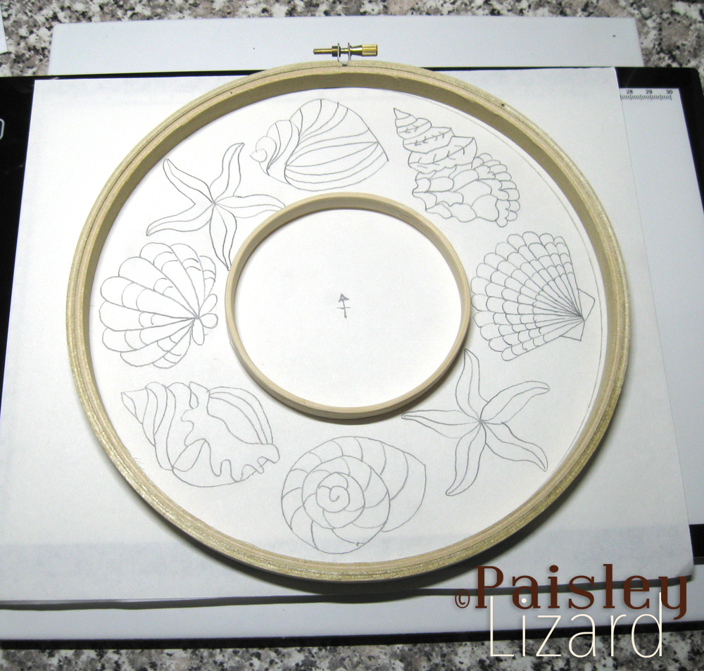 Two embroidery hoops laying on sketch of seashell wreath pattern.